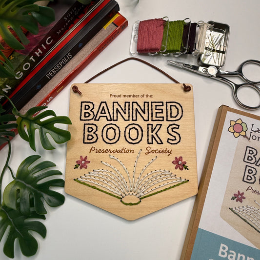Wooden banner embroidered with 'Proud member of the Banned Books Preservation Society' on a table with books and scissors.