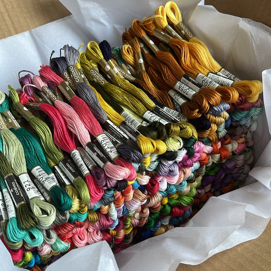 White tissue paper in a brown cardboard box holds a stack of 500 multicolored skeins of embroidery thread.