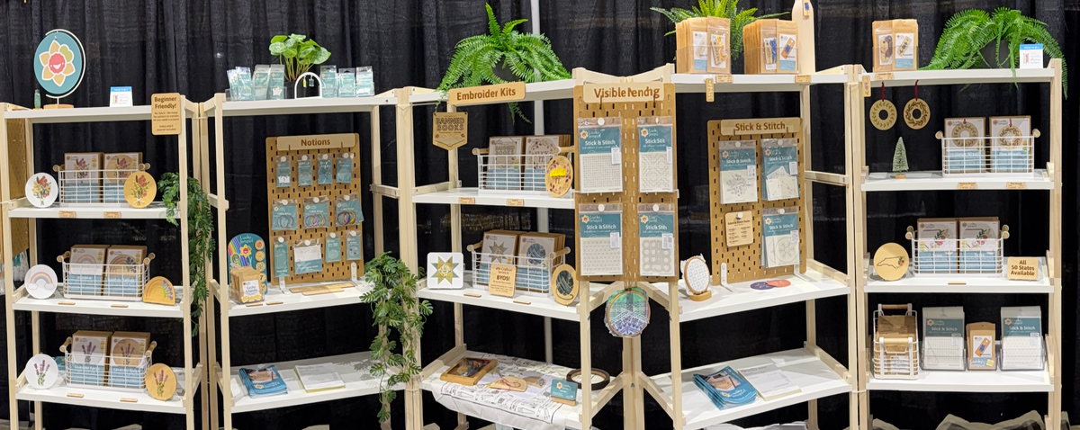 5 shelving units of pine wood and white shelves set up in Houston for the inaugural H&H Fall Trade Show set against black curtains. Shelves are filled with embroidery kits and samples, Stick & Stitch packets, shop catalog and order forms, and fake ferns and greenery.
