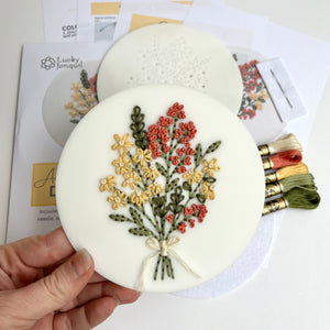 White acrylic disk with embroidery is held up, stitched with yellow daisies and pink flowers in a bouquet. Kit contents laid flat behind it.  