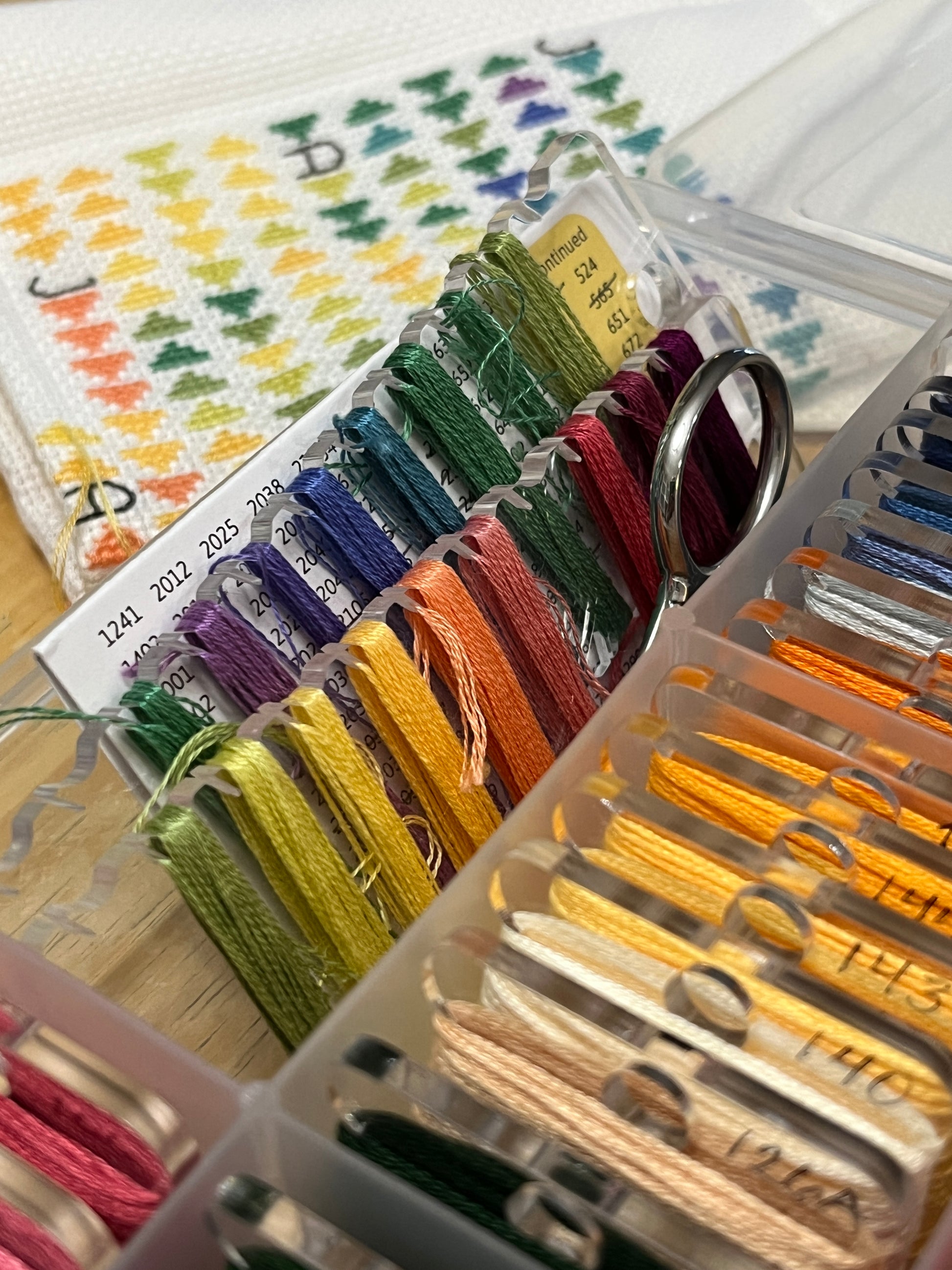 Closeup of two long floss keeps in a bobbin box with threads wrapped in rainbow order, other wound acrylic bobbins surround them in other spaces. A cross stitch project is in the background