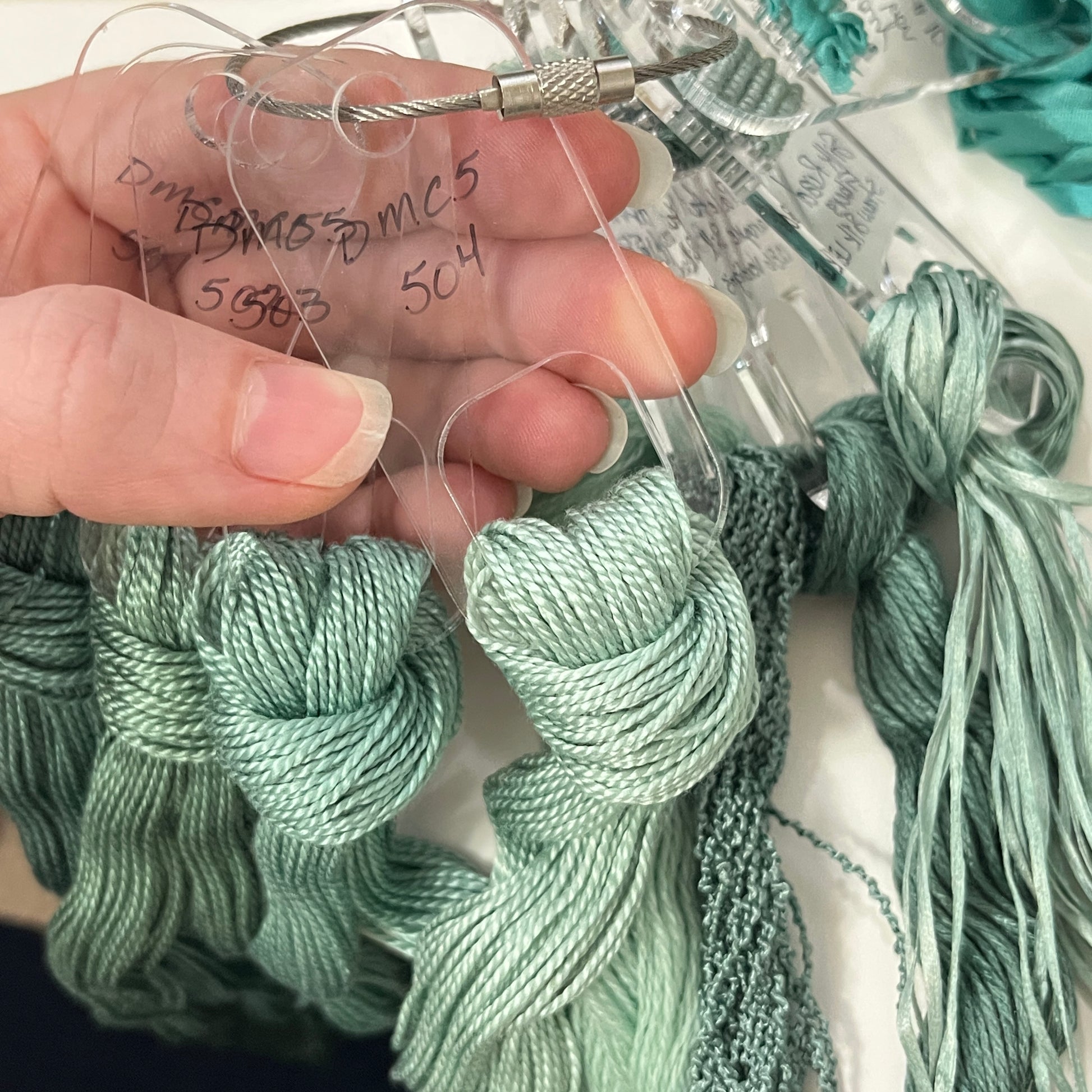 Clear acrylic needlepoint fiber drops on a cable ring with perle cotton in greens being held by a pale skin tone hand