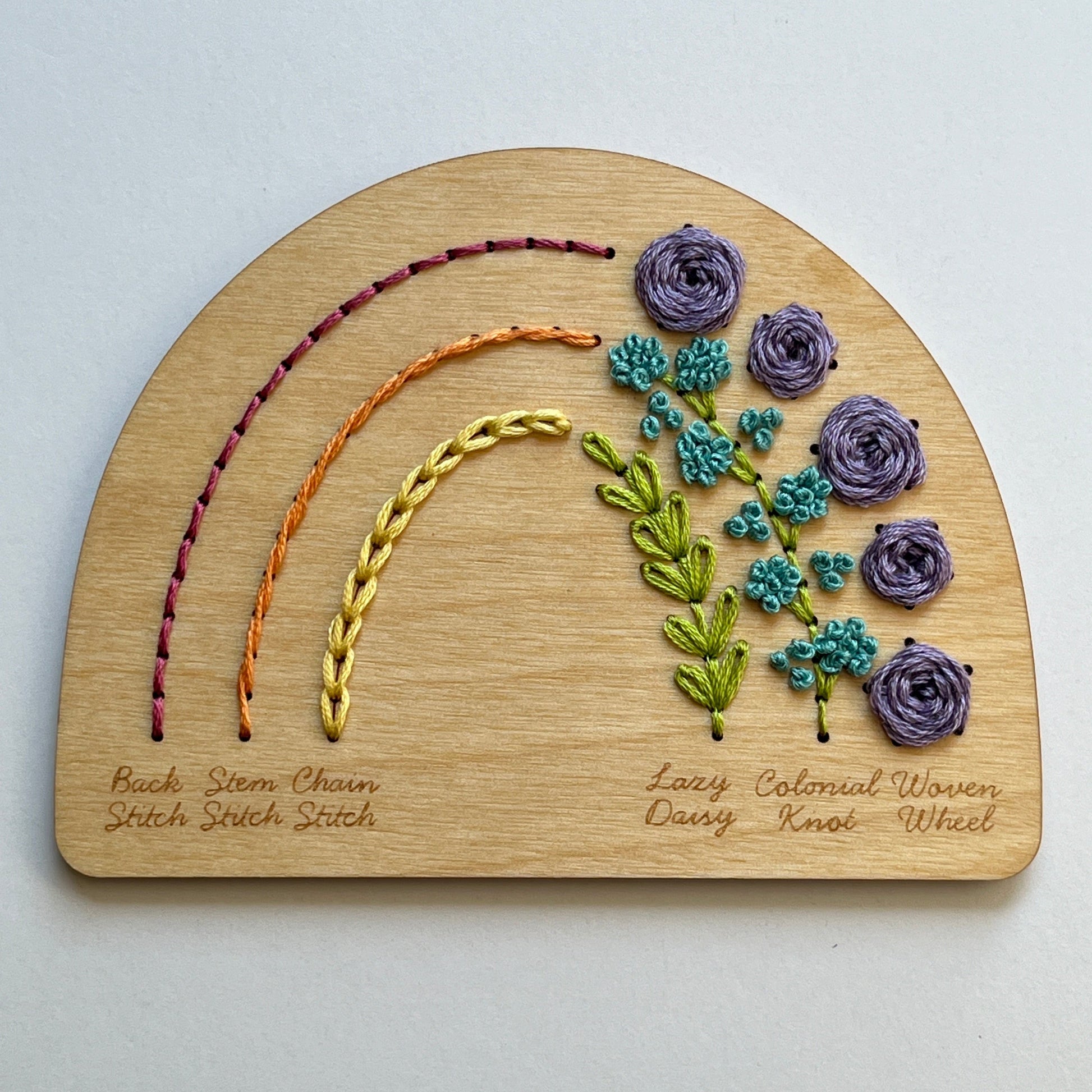 Wooden Laser cut plate in a half circle shape stitched with a colorful rainbow of floss illustrating six different labeled stitches